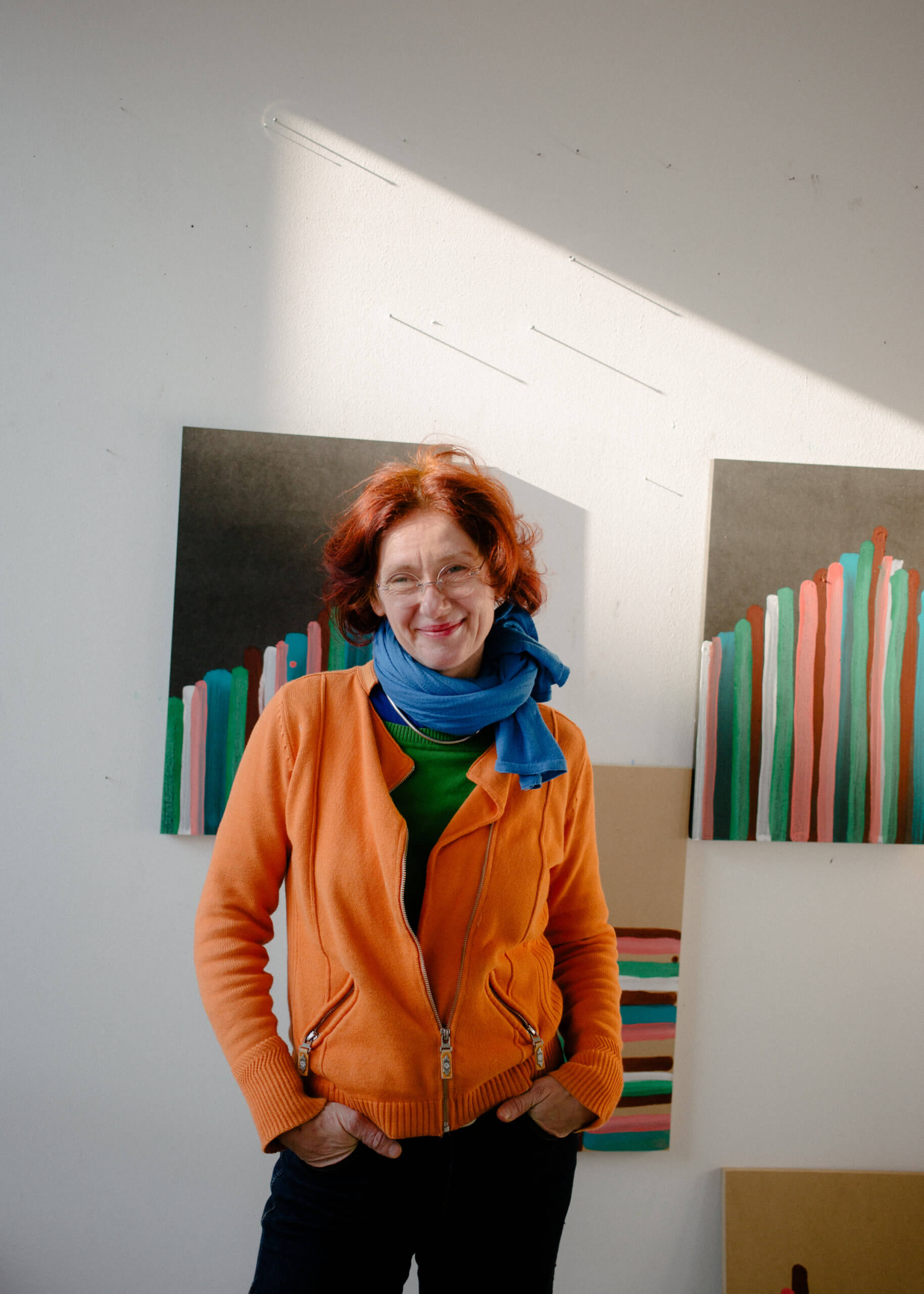 Ein Portrait von Annegret Hoch in ihrem Atelier. Sie steht vor einer weißen Wand an denen unfertige Arbeiten von ihr hängen.